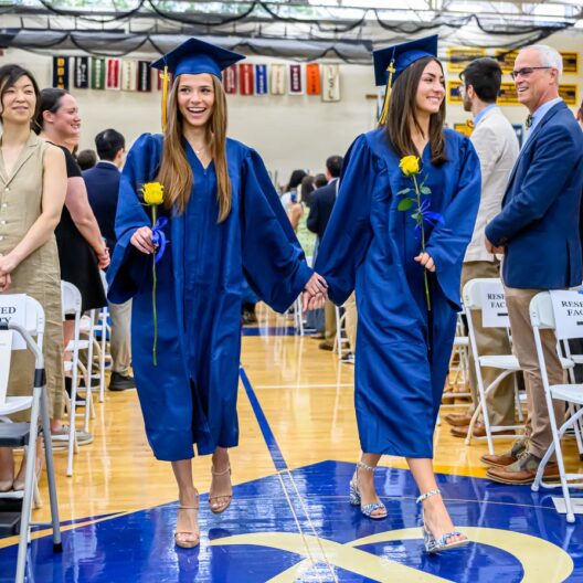 Two graduates wearing blue caps and gowns walk hand-in-hand down an aisle, holding yellow flowers during a graduation ceremony.