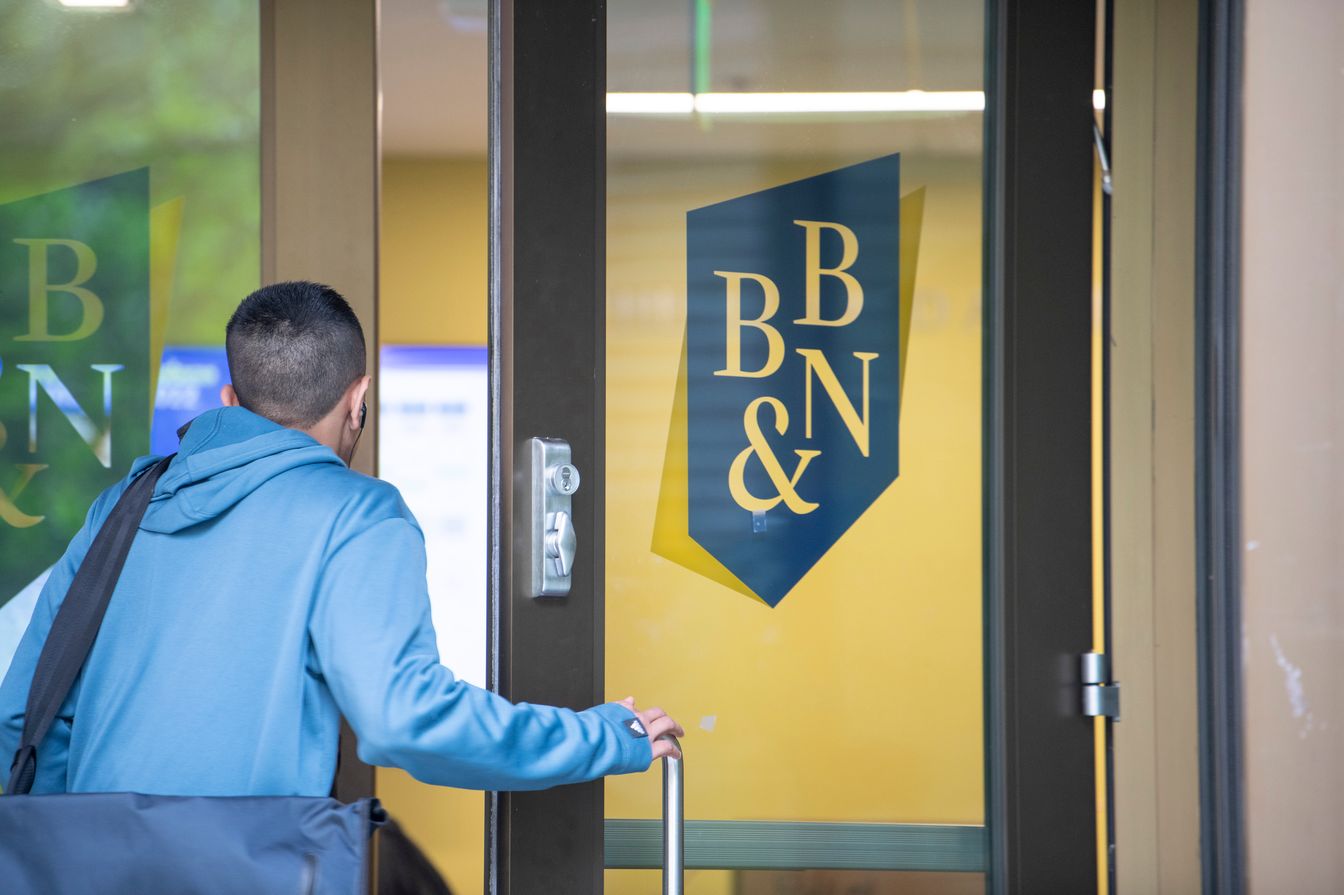 A person in a blue hoodie and carrying a bag opens a glass door with the BB&N logo on it.