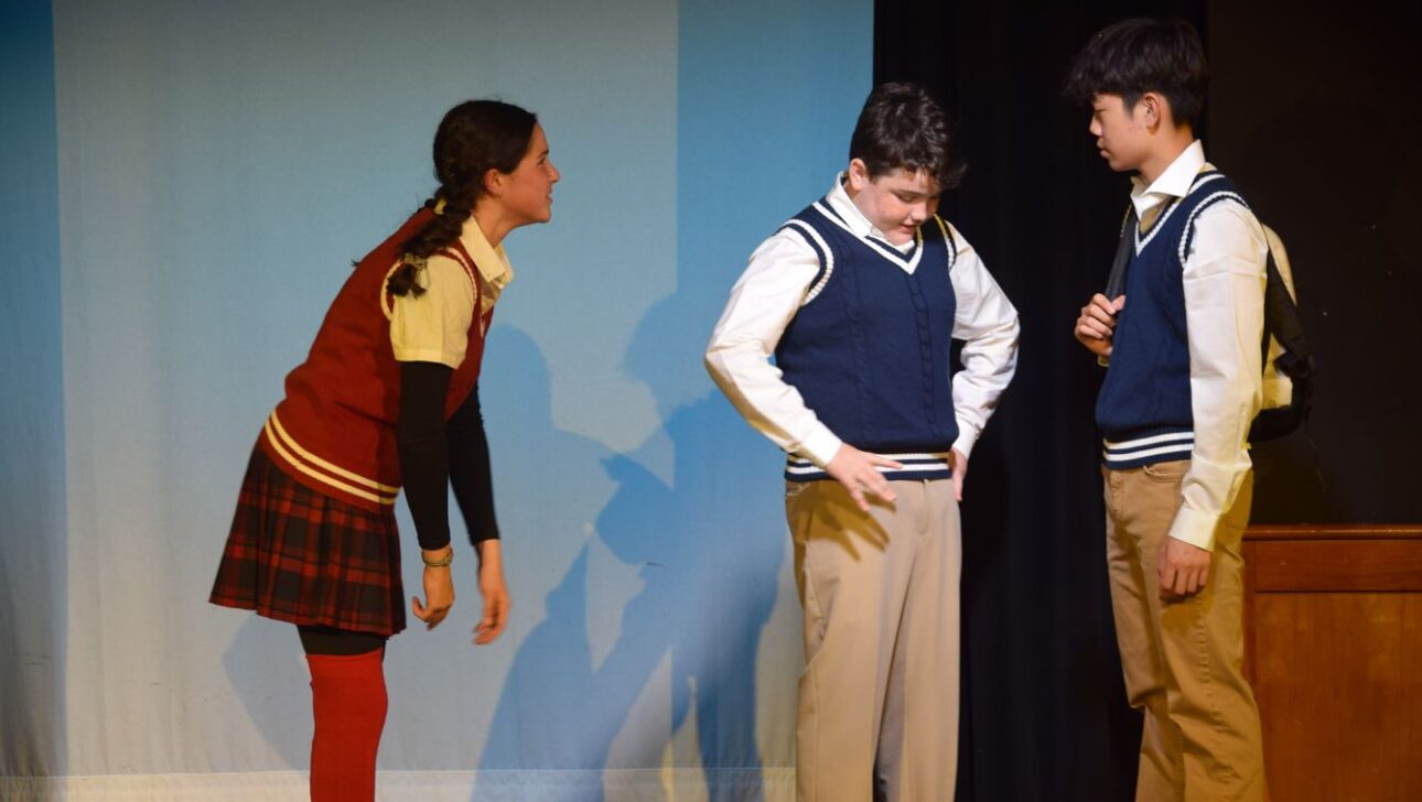 Three young actors perform on stage, wearing school uniforms.