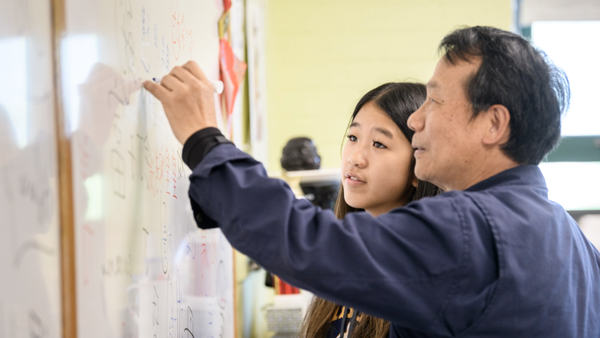 A teacher instructs a student at a whiteboard, writing mathematical equations. The teacher is pointing at the board, and both are focused on the lesson.