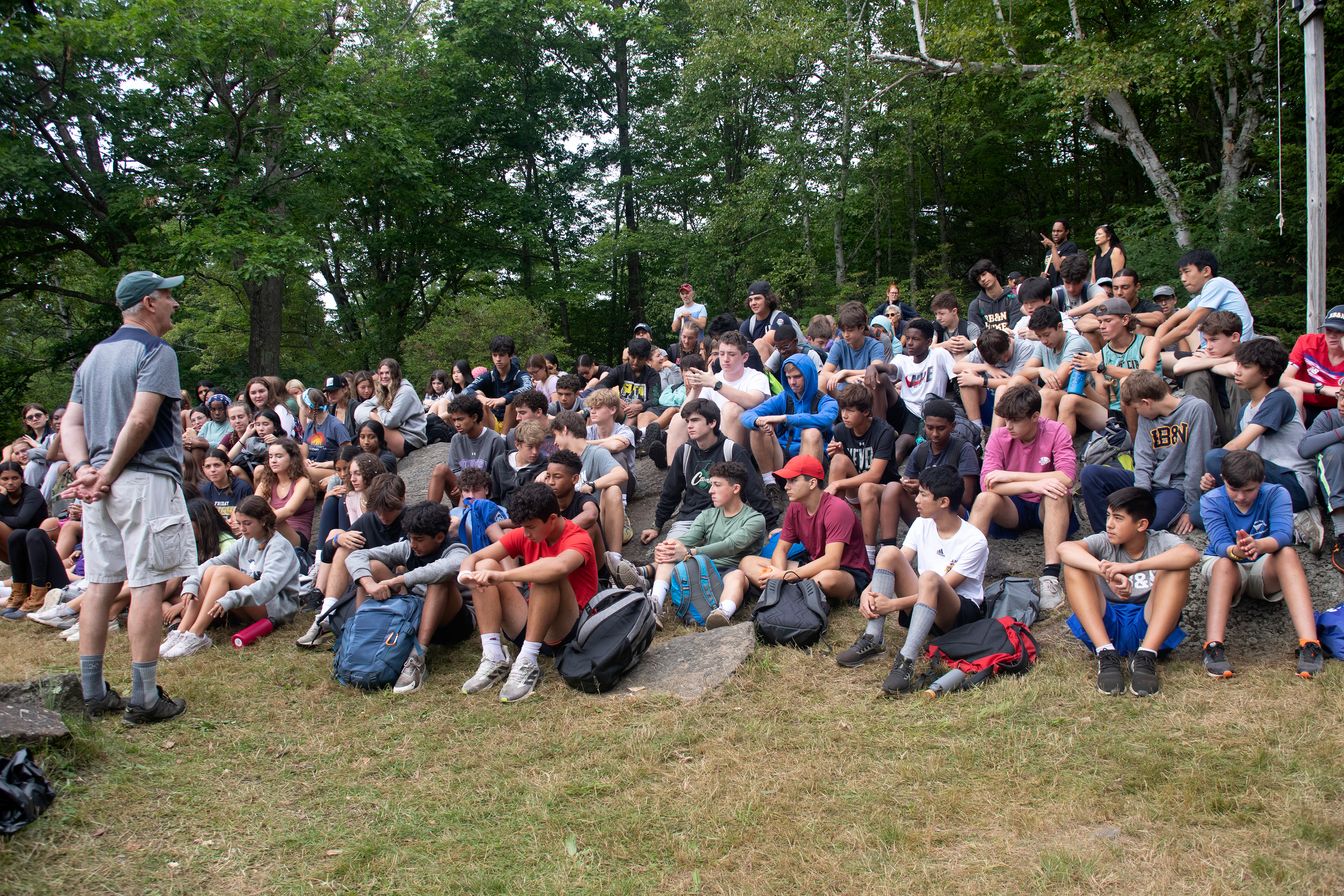 A group of people, predominantly students, sits on outdoor steps and grass, attentively listening to a person standing and speaking.
