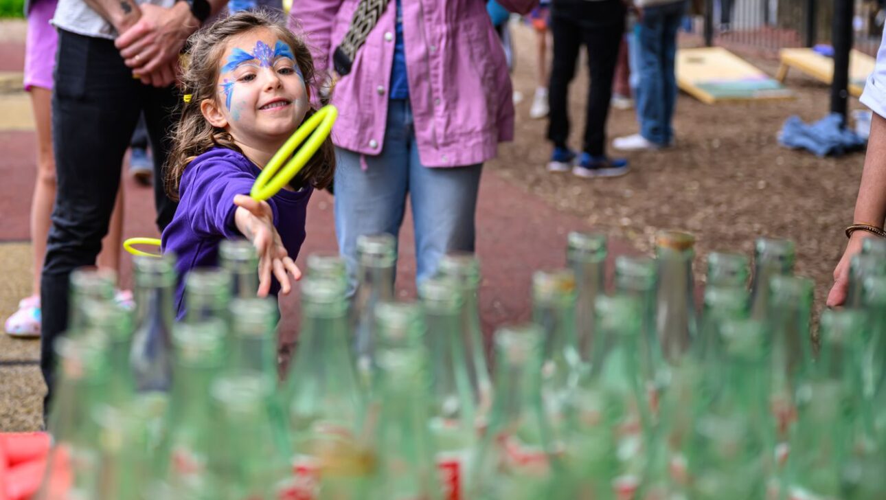 A young girl with face paint throws a ring toward glass bottles at a carnival game while several people stand in the background.