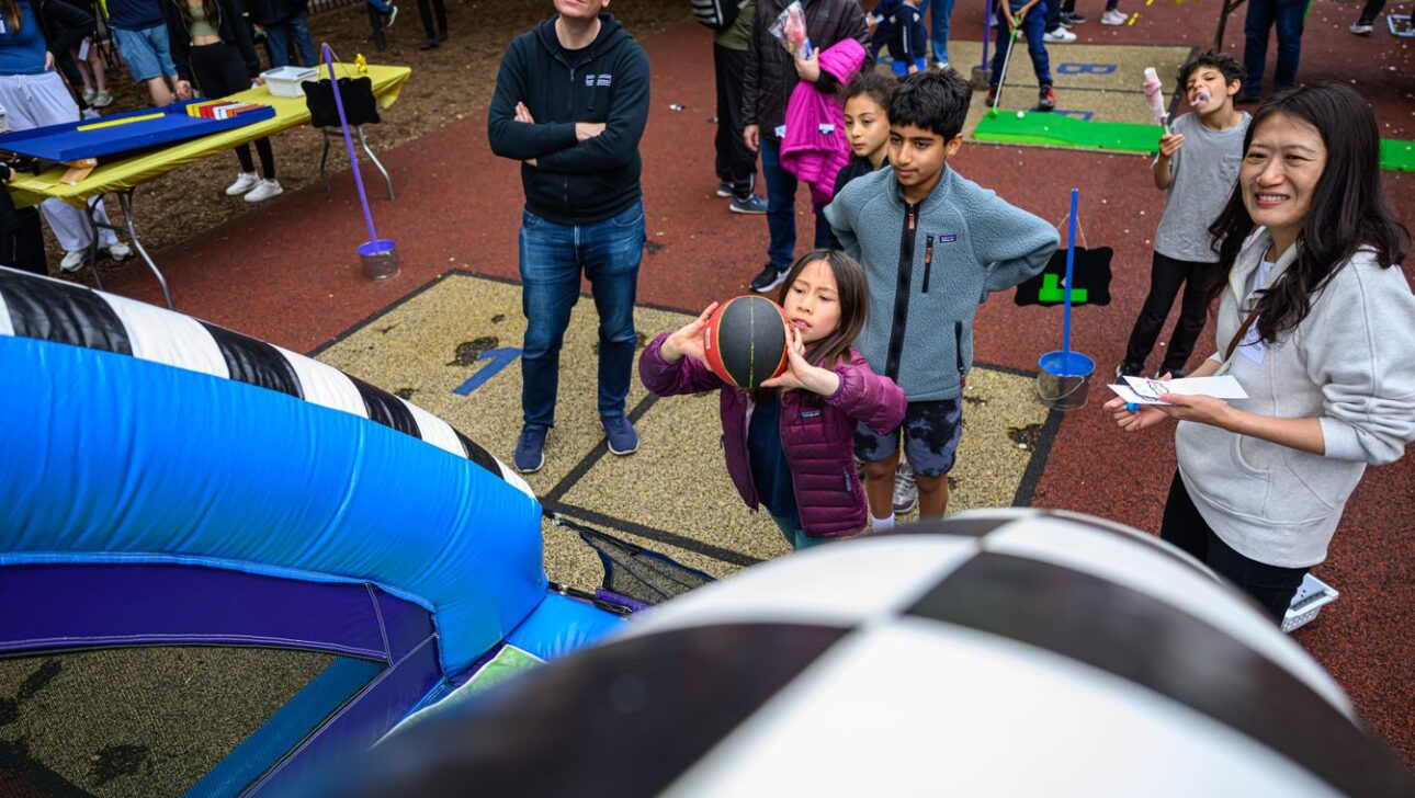A young girl prepares to shoot a basketball at an inflatable hoop while others look on at an outdoor event.