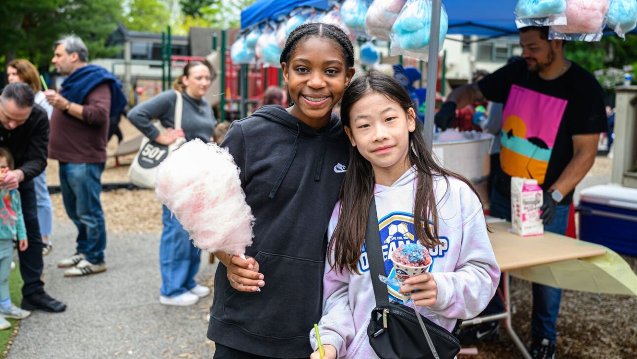 Two girls standing side by side at an outdoor fair, one holding a cotton candy and the other with a small treat. Other people and a booth with cotton candy can be seen in the background.