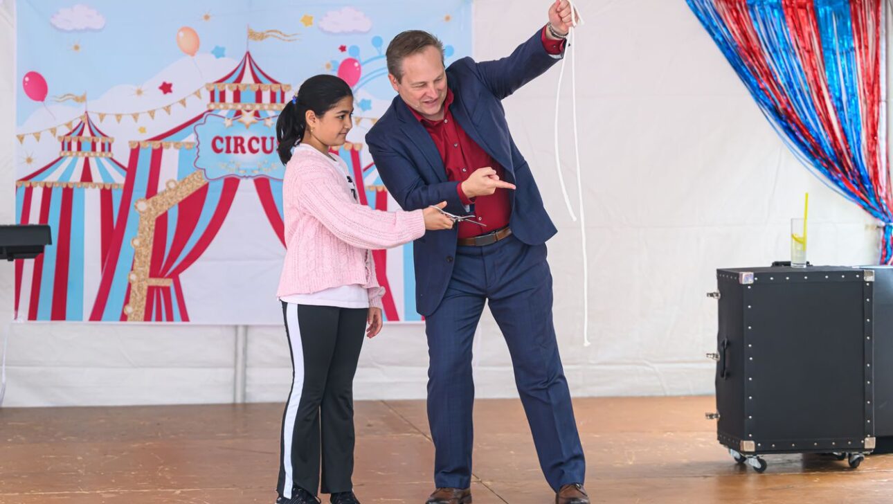 A magician and a young girl perform a rope trick on a stage decorated with circus-themed banners. The magician points at the rope, while the girl holds part of it, both focused on the trick.