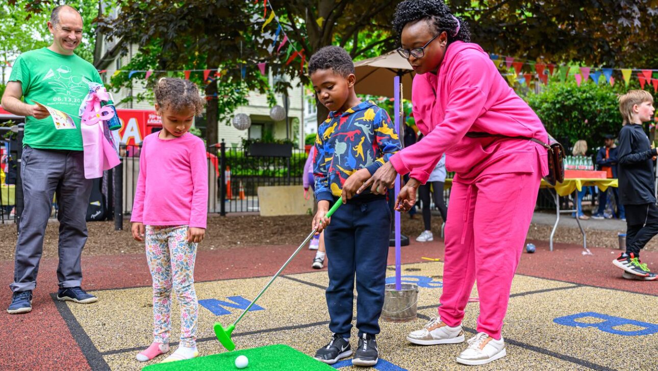 An adult assists a child in putting a golf ball while another child stands nearby on a colorful play area outdoors. A man observes the scene from the side, holding a flyer.