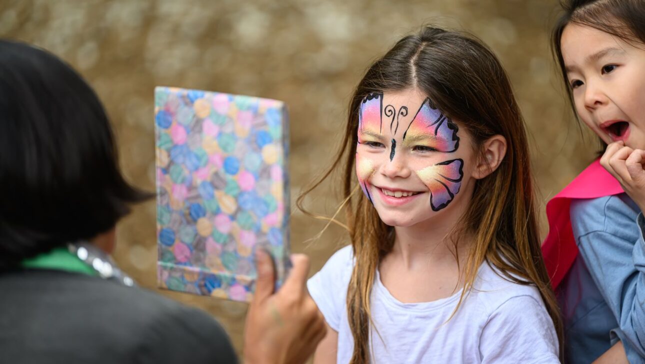 Child with butterfly face paint smiles while looking at a mirror held by an adult. Another child stands beside with an open-mouthed excited expression.