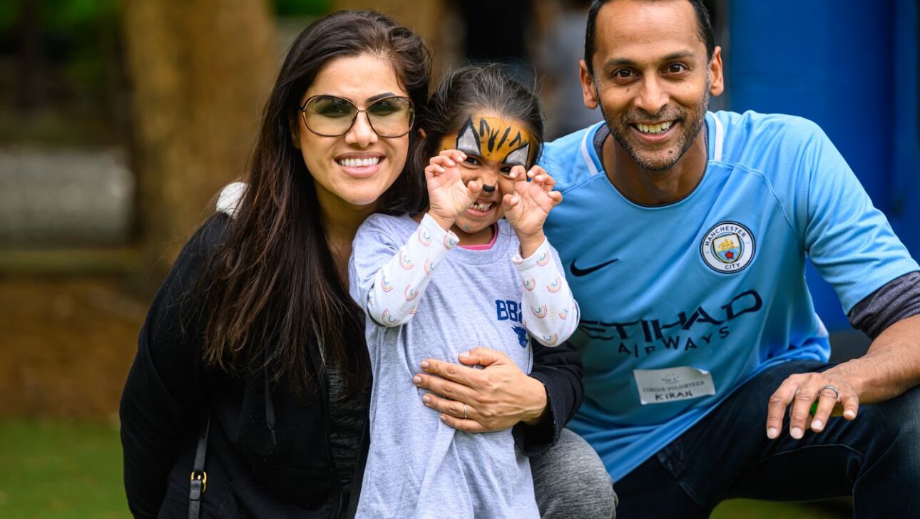 A man in a sports jersey and woman in sunglasses pose with a child who has face paint, outdoors.