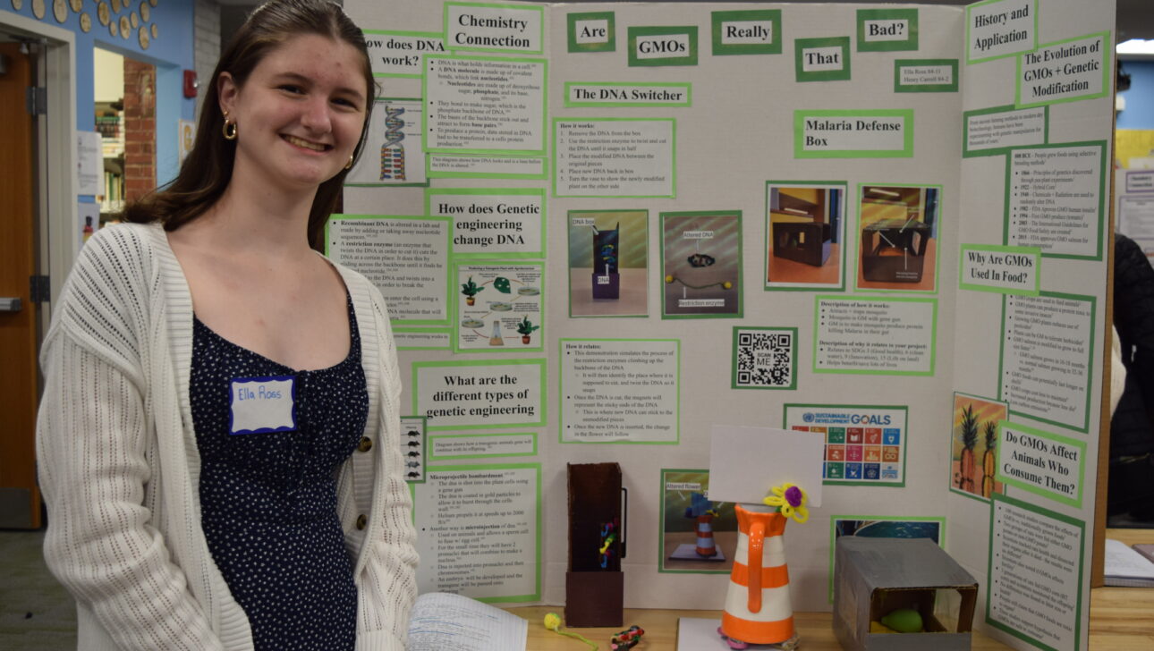 A young person stands next to a science fair project display board featuring information on genetic engineering, DNA, and GMOs.