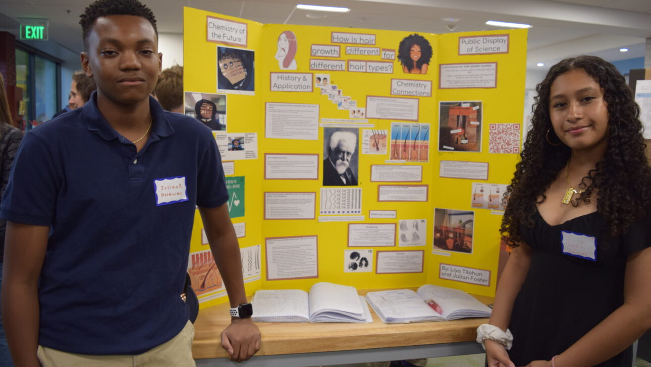 Two students stand beside a yellow tri-fold presentation display board in a brightly lit room. The board features various images, texts, and diagrams.