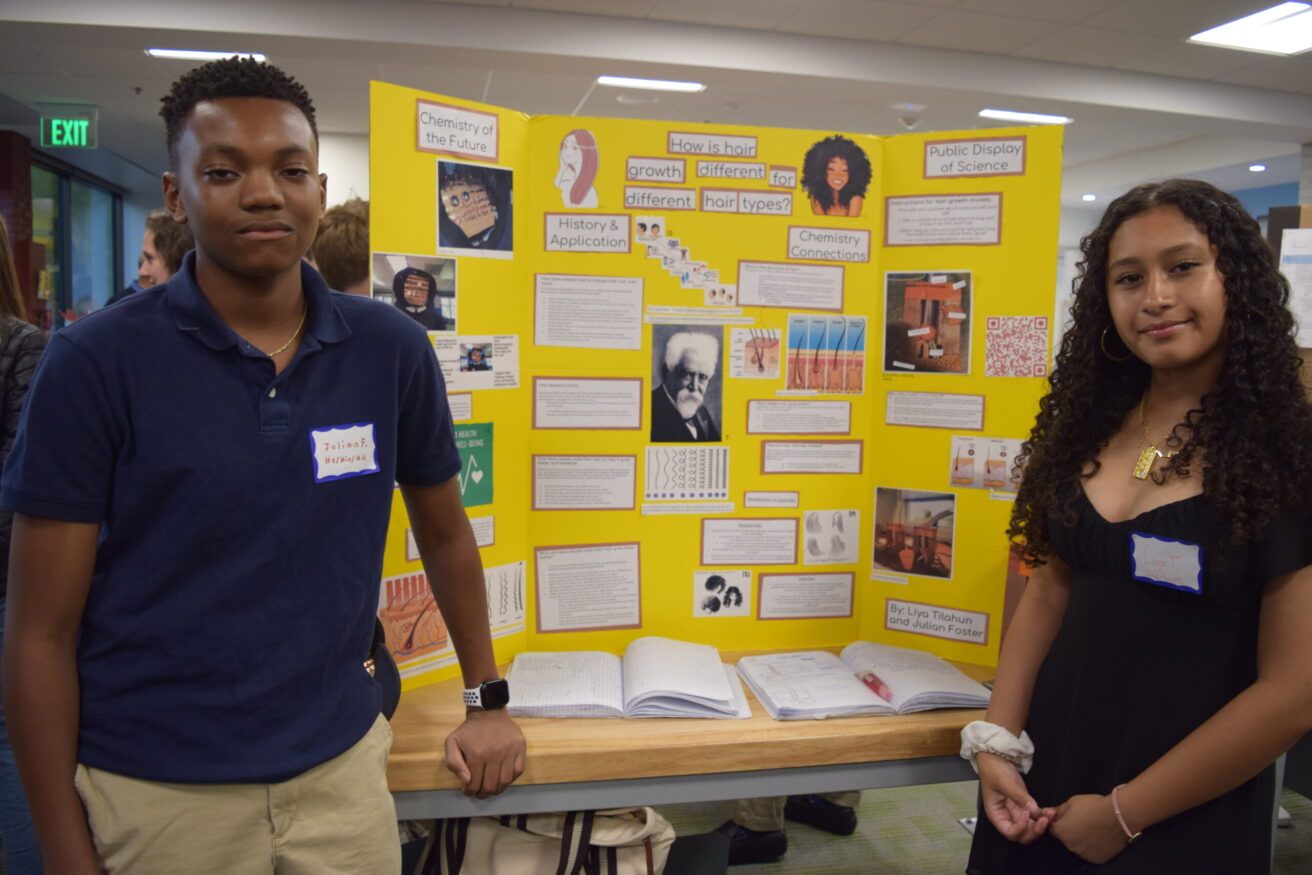 Two students stand beside a yellow tri-fold presentation display board in a brightly lit room. The board features various images, texts, and diagrams.
