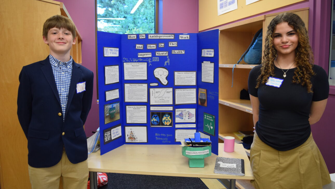 Two students stand beside their science project display titled "How Antidepressant Work and How They Improve Mental Health," which features text, diagrams, and images explaining their research.