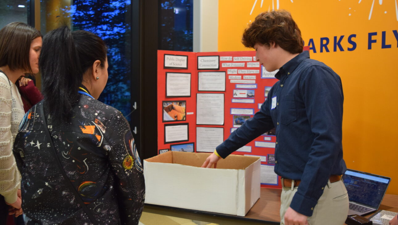 A young man presents a science project on a display board to two women.