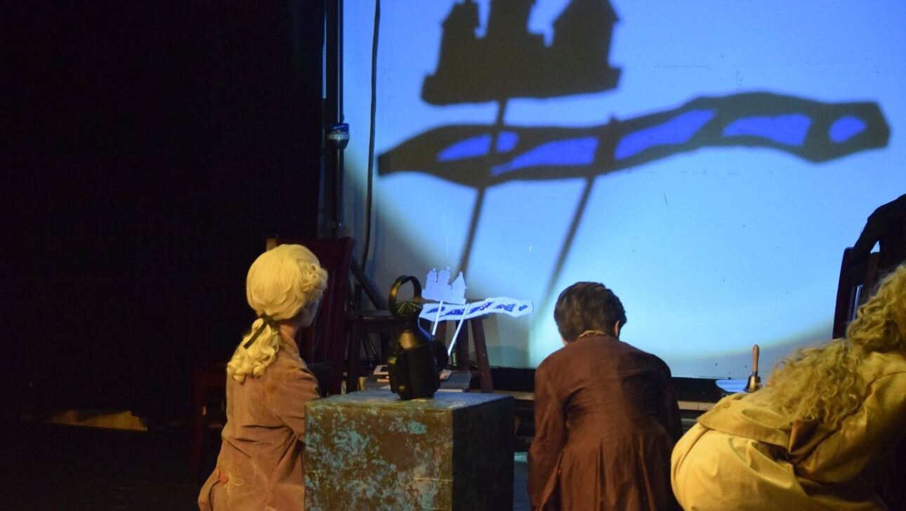 Three individuals are watching shadow puppets of a house and a tree being projected onto a screen in a dark room.
