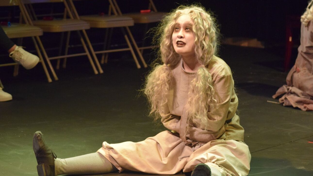 A person with long, curly hair, wearing a beige costume and stage makeup, sits on the floor with legs apart and arms folded in front, performing in a theater setting with chairs in the background.