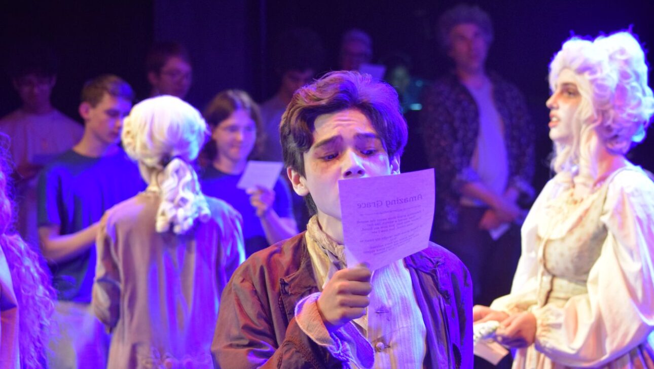 A young man in period costume reads a paper in a dimly lit room with other similarly dressed individuals and onlookers in the background.