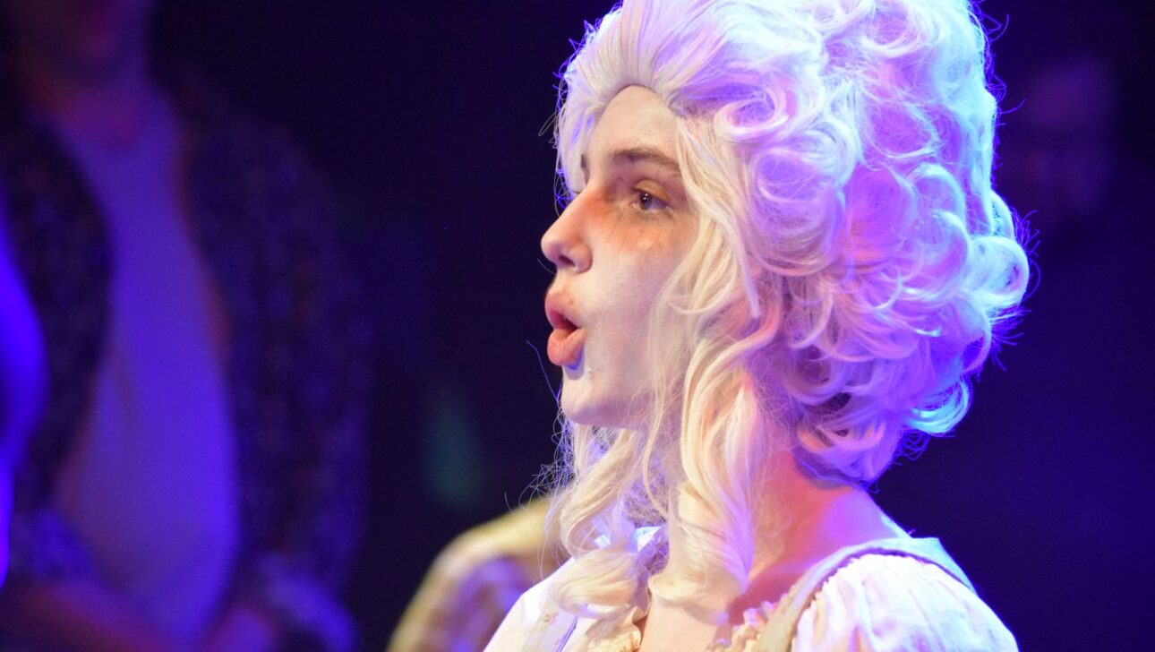 A person in colonial-era costume with a white wig and face makeup performs under stage lighting.
