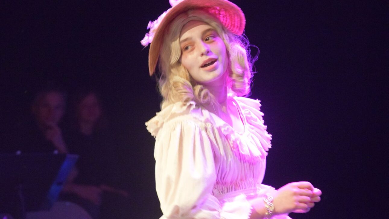 A person with curly hair, dressed in 18th-century style attire and a hat, is illuminated by stage lights while performing.
