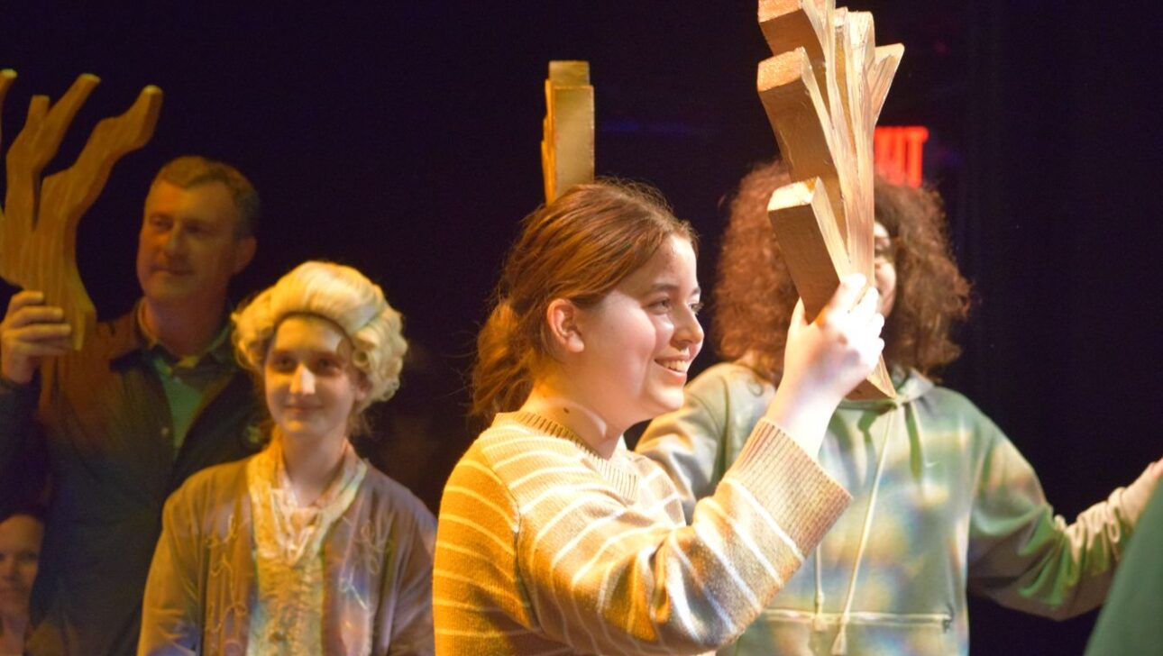 A group of people, including a smiling girl in the foreground, are holding wooden shapes above their heads in a dimly lit setting.