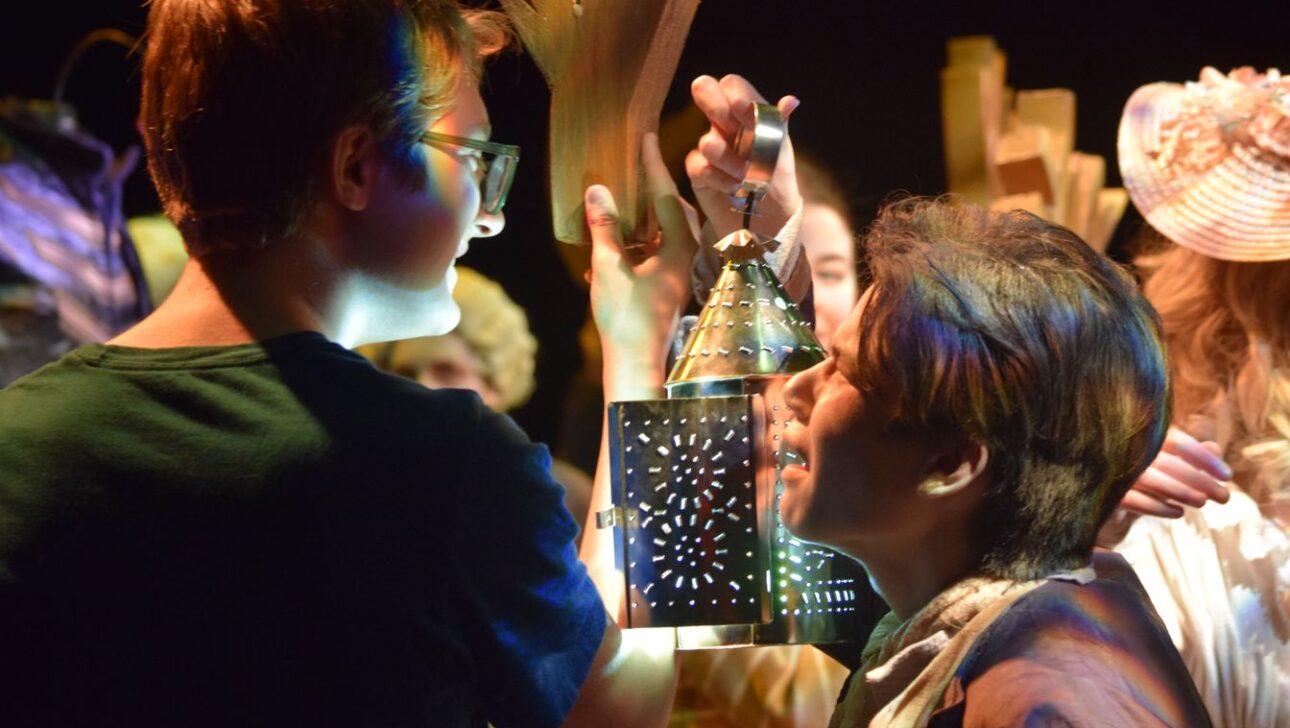 Two people in costume, one holding a decorative lantern with a candle, providing light. The scene appears to be part of a theatrical performance with other people in background.