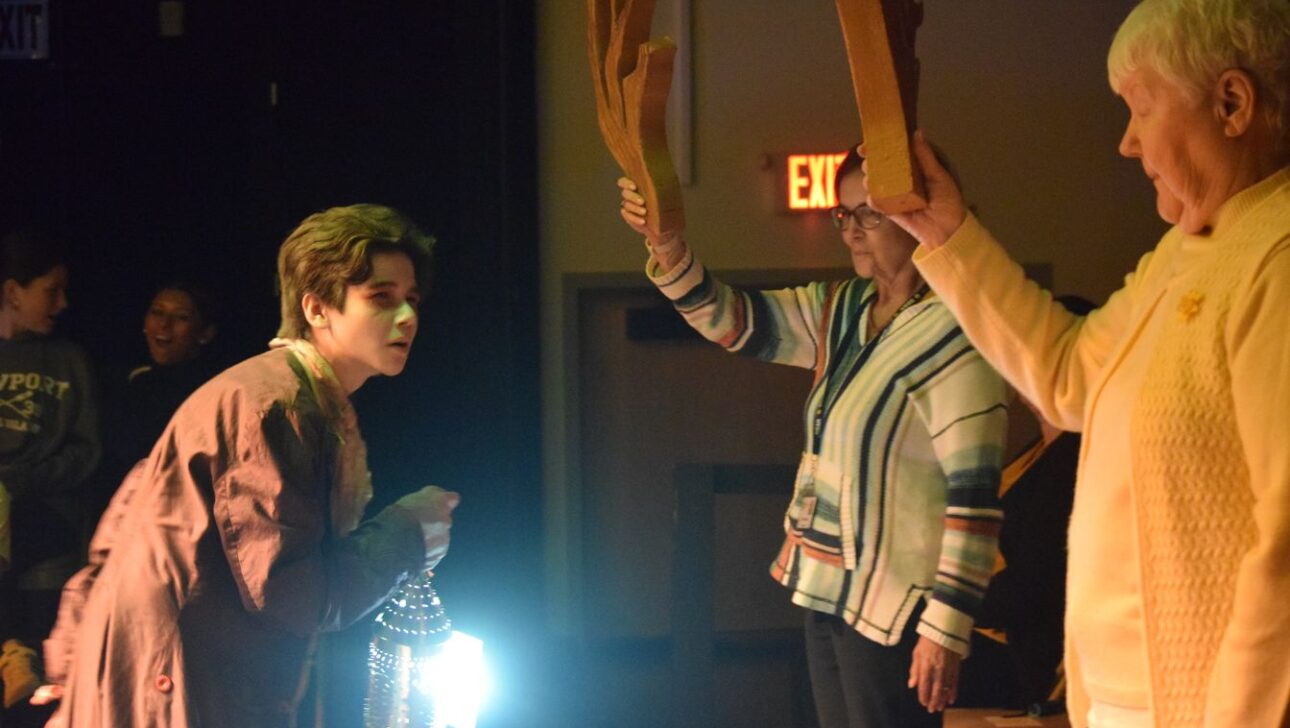 A person holds a lantern and leans forward on a dim stage, while two others raise wooden tree branches in the background.