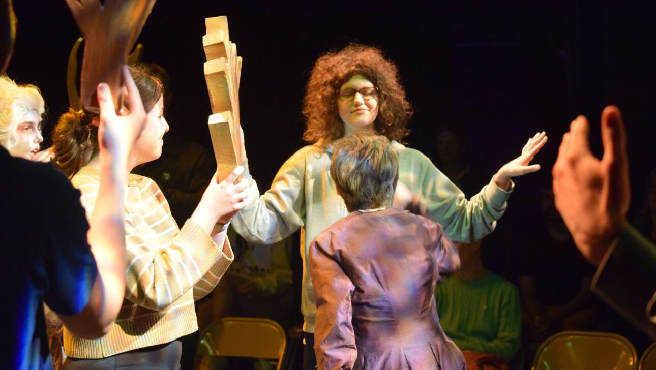 A group of people in a dimly lit room, one person holds wooden objects up while another, wearing glasses and curly hair, seems to be making a gesture. Chairs and some seated individuals are in the background.