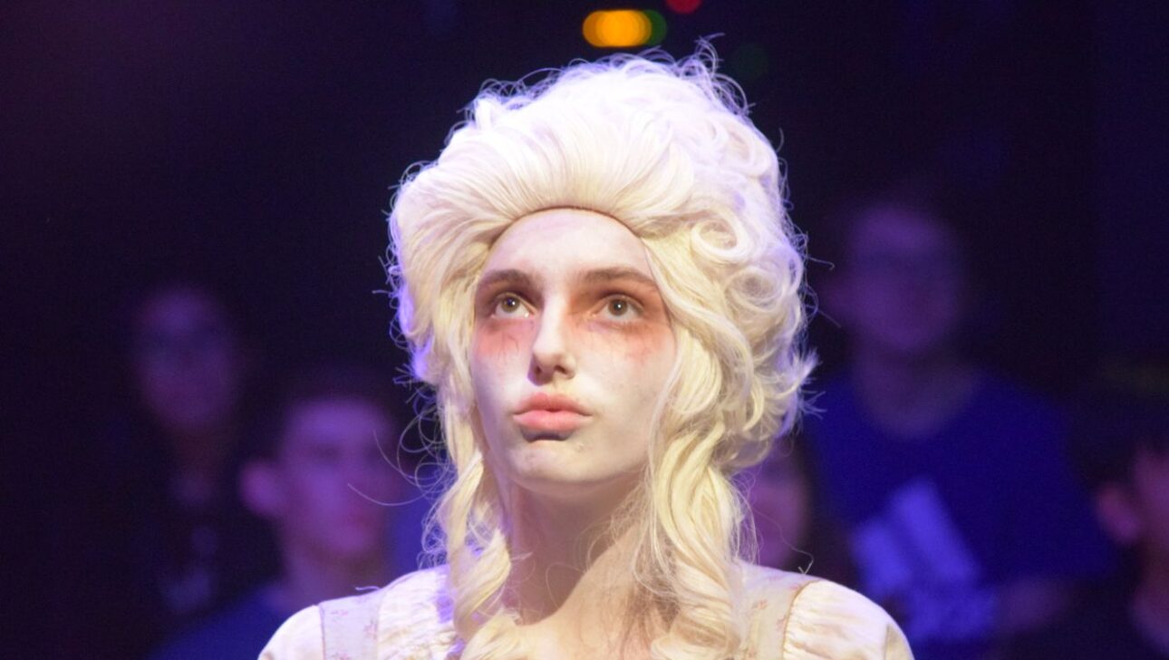 A person dressed in 18th-century style costume with a white wig and pale makeup is illuminated under stage lighting, with people in the dim background.