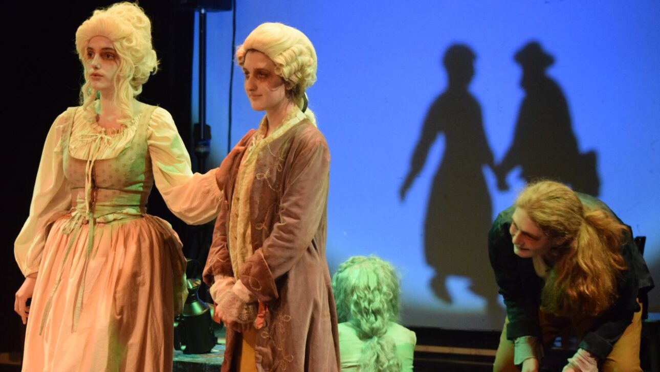 Four people in vintage costumes stand on stage with a blue backdrop. Two shadows are visible on the backdrop. One person sits on the floor, and another kneels while two interact in the foreground.