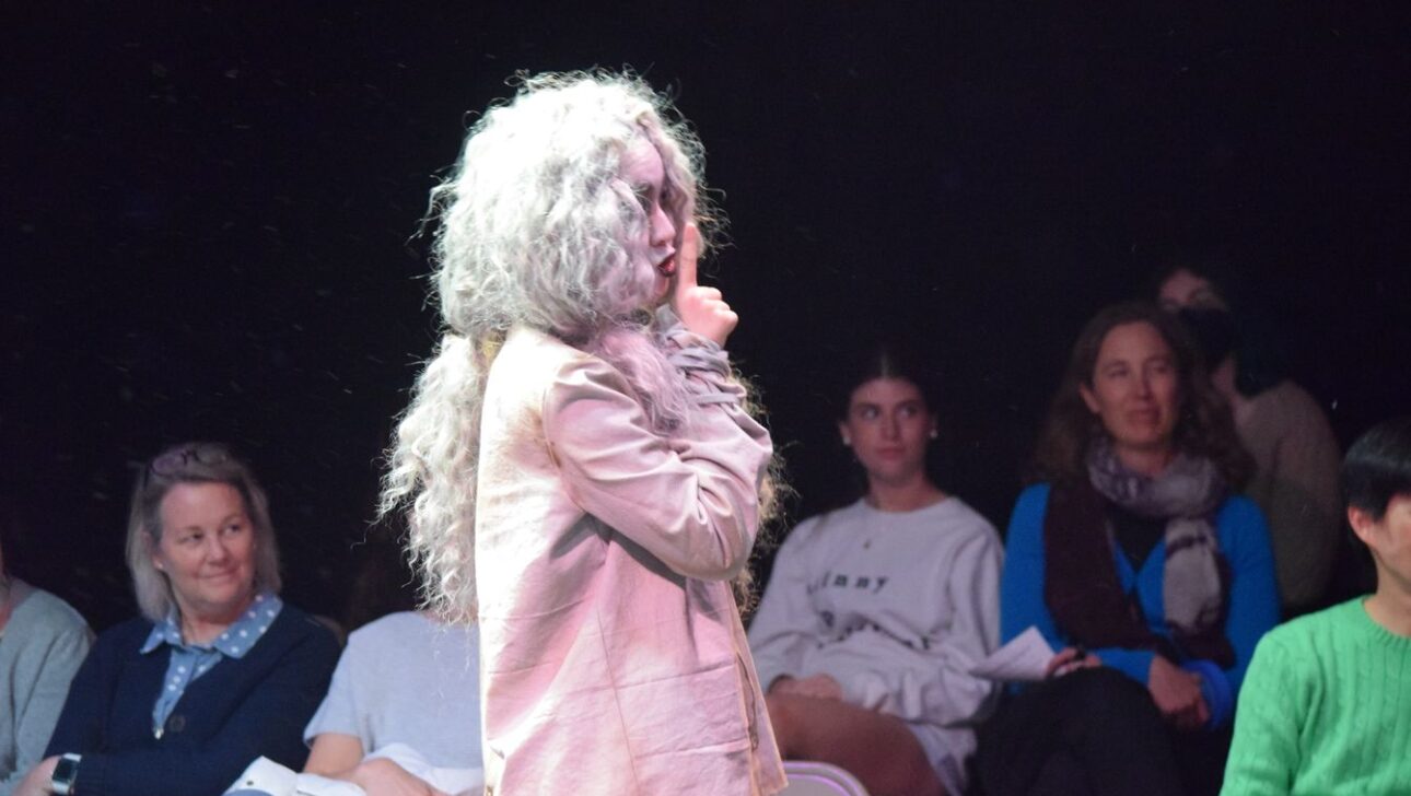 Person with long, gray, messy hair and light-colored clothing stands on a stage with a finger raised to their lips, suggesting silence. Seated audience members observe attentively in the background.