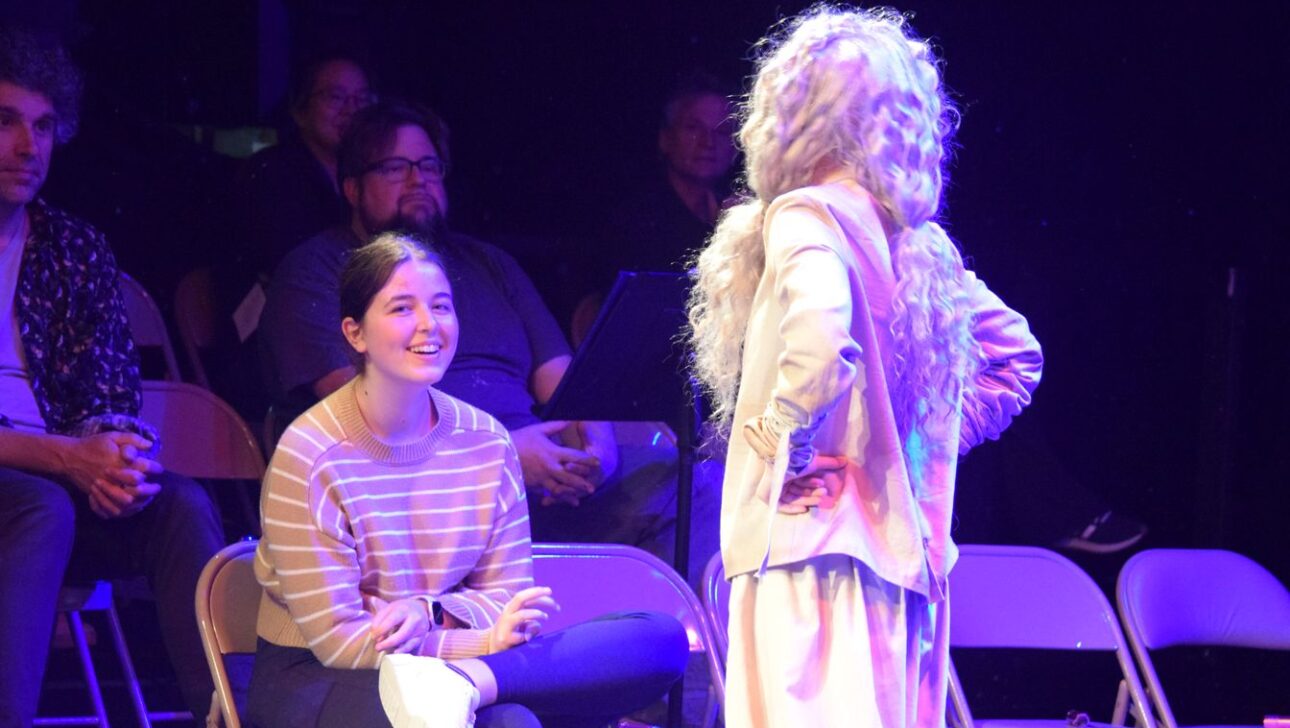 A person with long, light-colored hair stands facing another person who is sitting and smiling. Several people are seated and observing in the background.