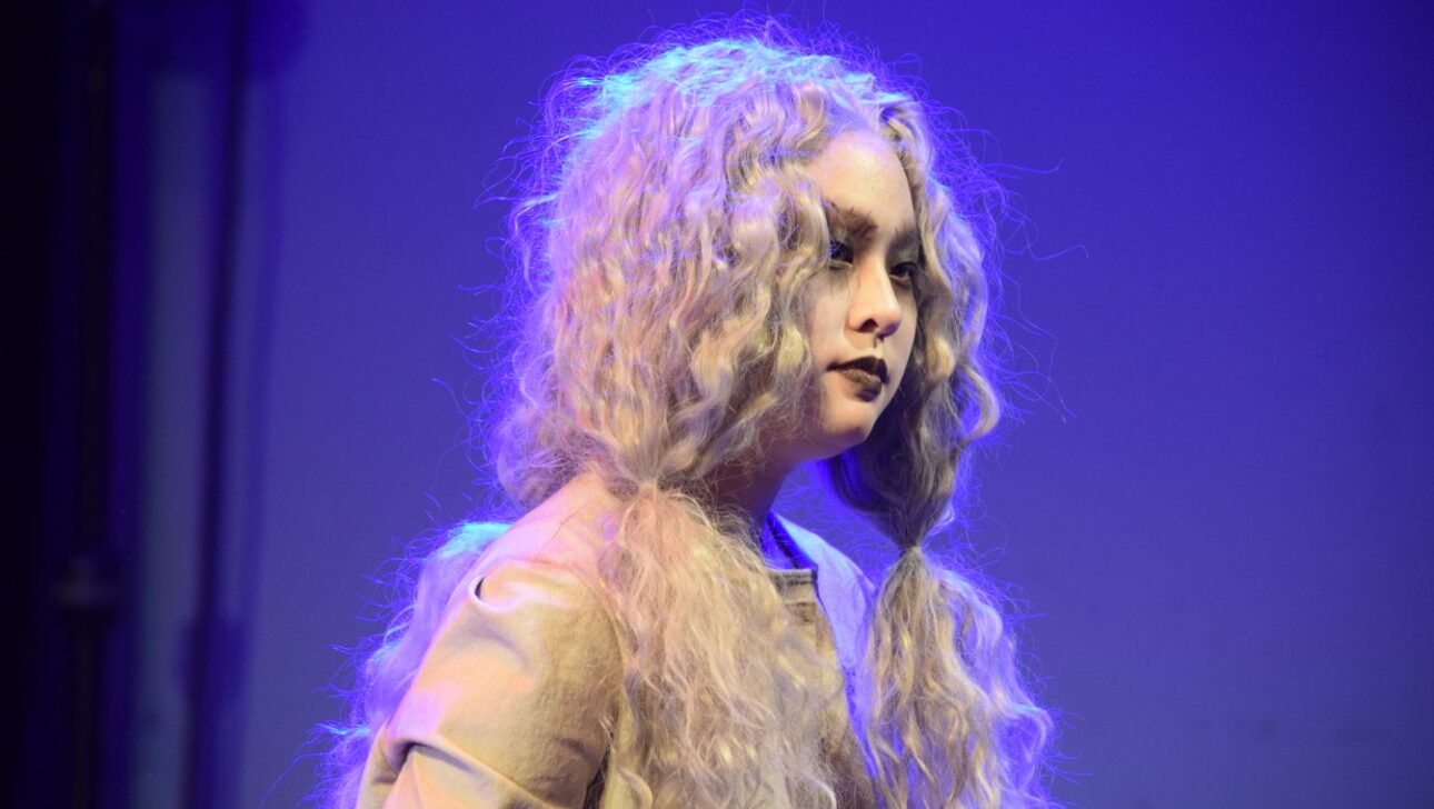 Person with long, curly, silver hair stands against a blue-lit background, dressed in a neutral-colored outfit.