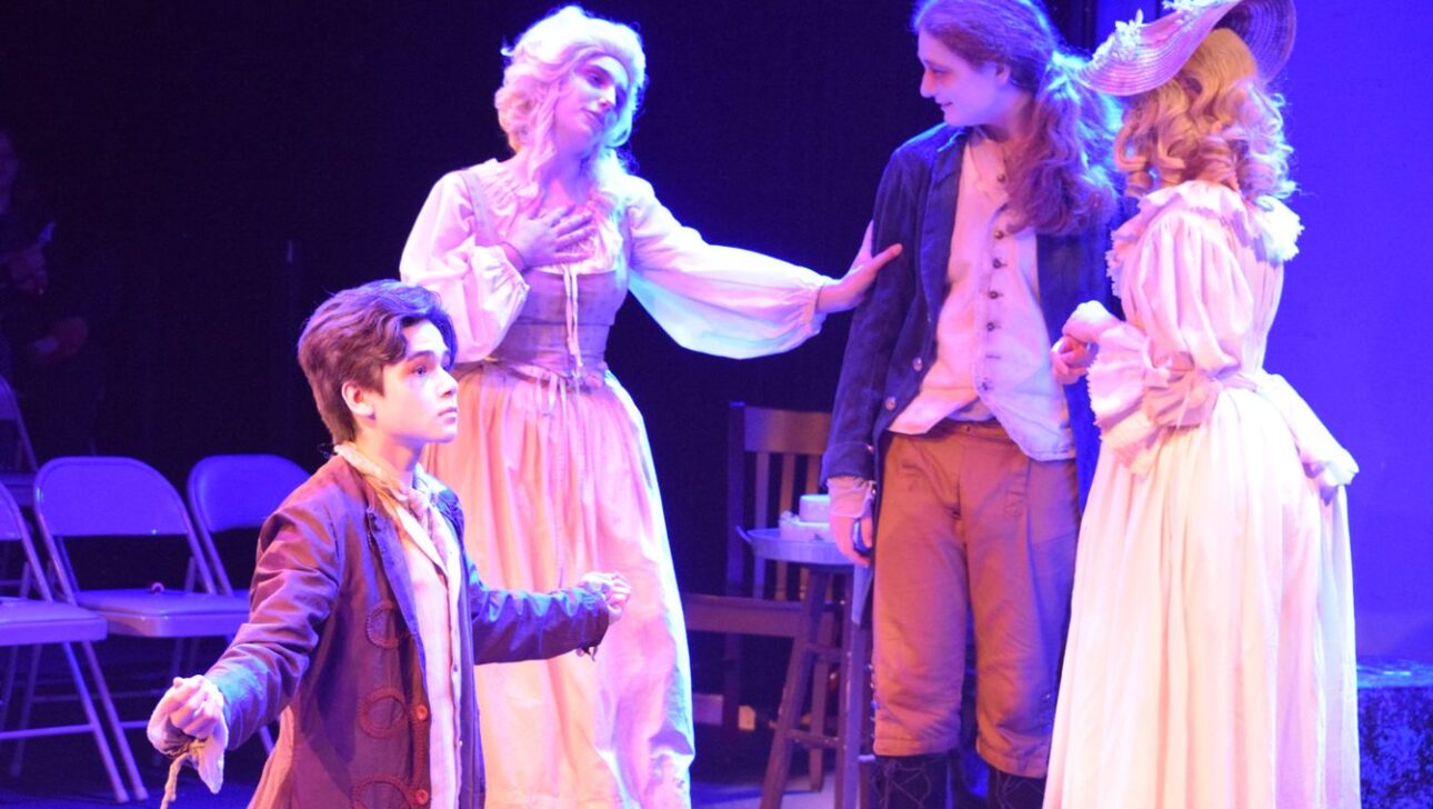 Four actors in period costumes perform a scene on stage. One actor gestures expressively in the foreground, while three others interact in the background. .