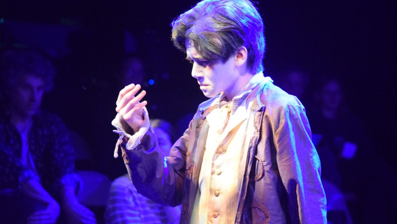 Person in period costume, illuminated by stage lights, gazes intently at their hand. Audience members in the background watch attentively.