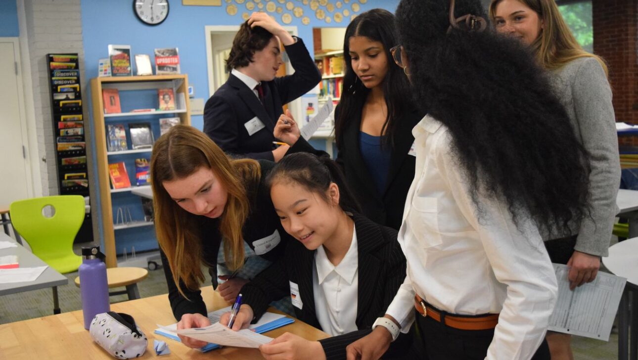 A group of students, both standing and sitting, are engaged in conversation and reviewing documents at a table in a classroom or library setting.