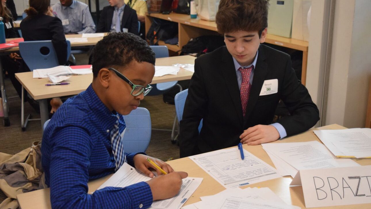 Two students in formal attire are seated at a classroom table, working on documents labeled "Brazil."