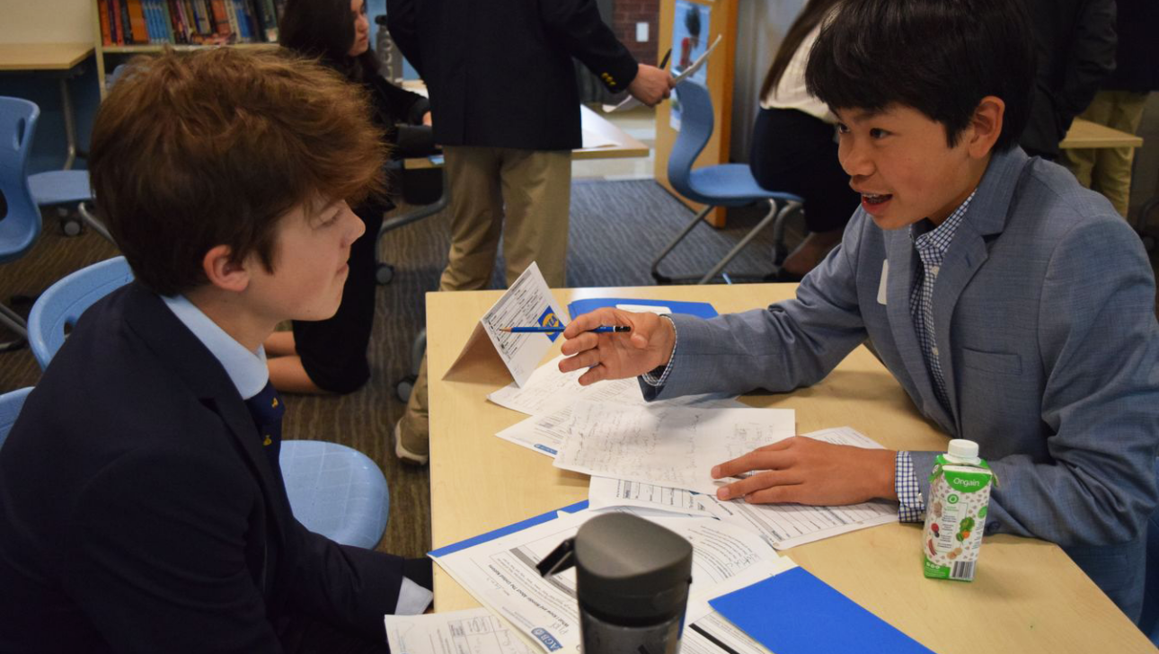 Two boys in school uniforms sit at a table engaging in a discussion, with papers and a box of organic milk in front of them.