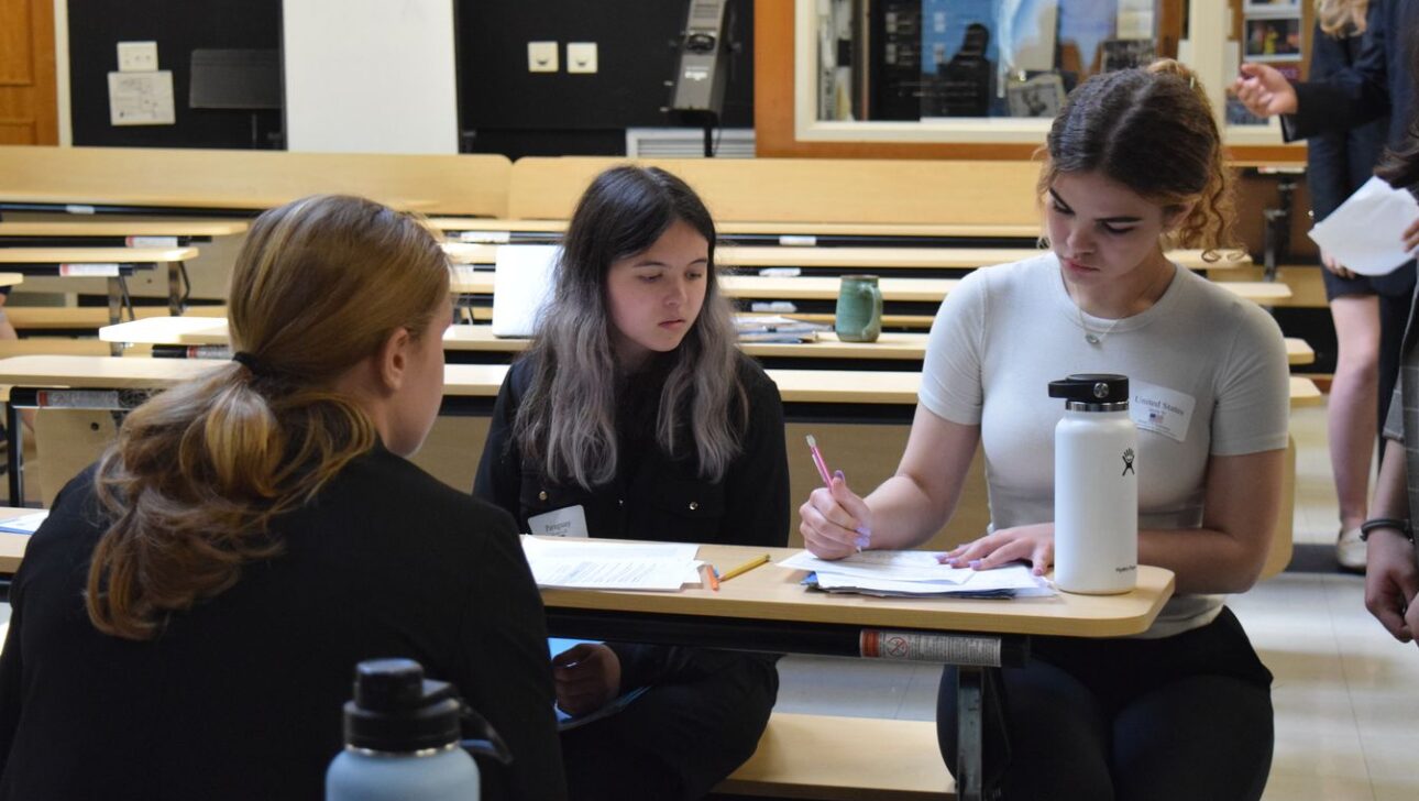 Three individuals are seated around a table in a classroom, focusing on paperwork.