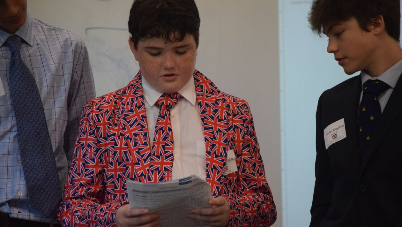 A boy in a suit patterned with the Union Jack is reading from a paper.