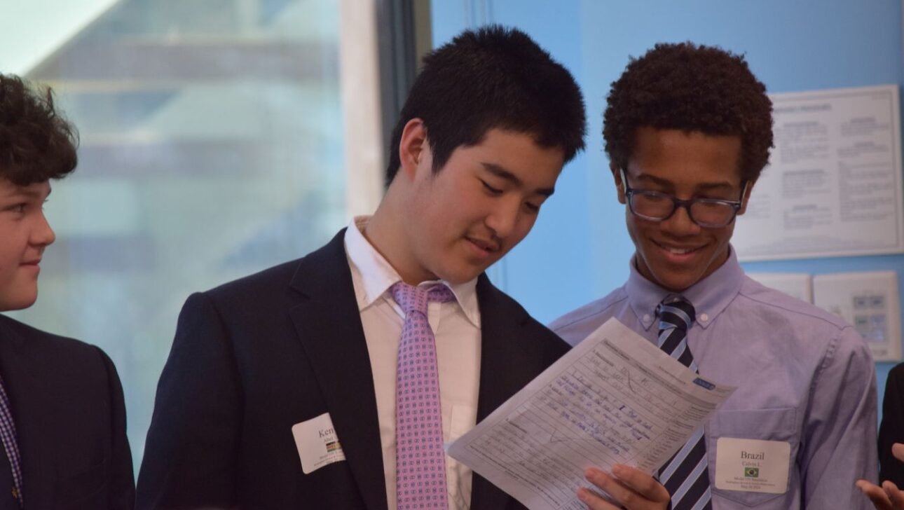 Three young men in formal attire review a document together, standing in a brightly lit room with windows.