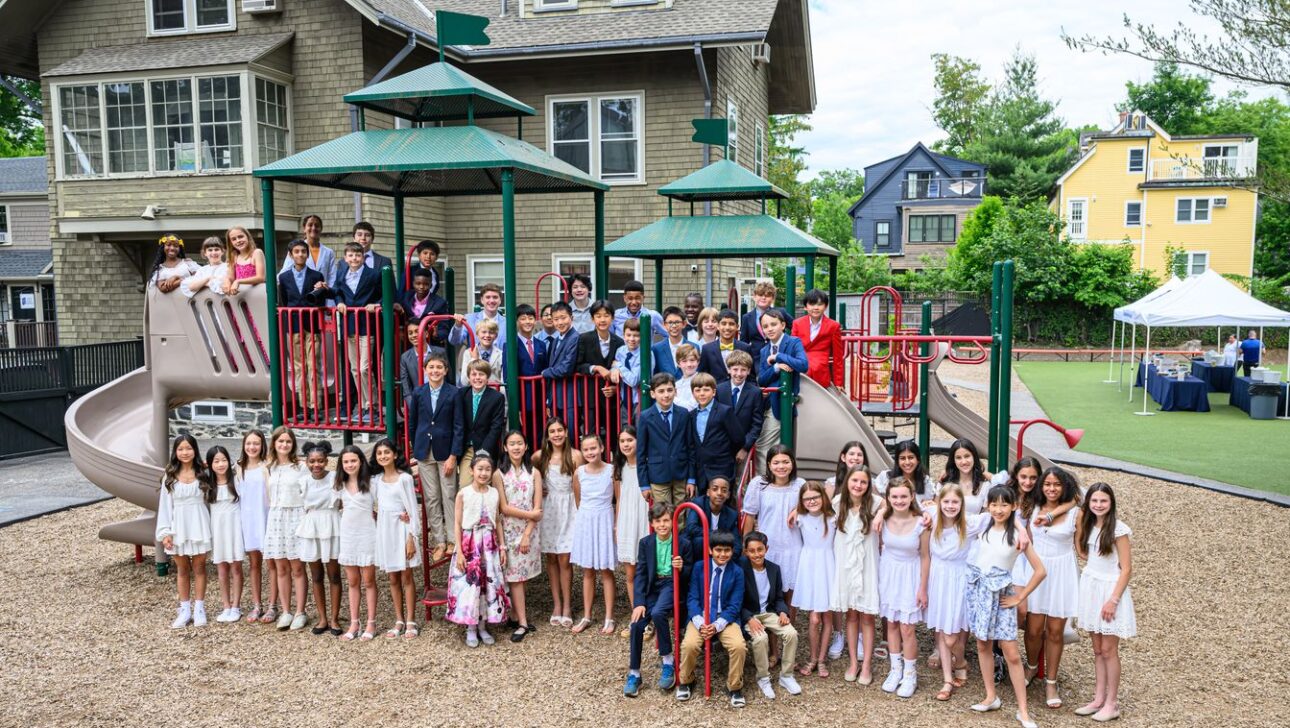 A large group of children, mostly dressed in white, poses on playground equipment in front of a residential-style building.