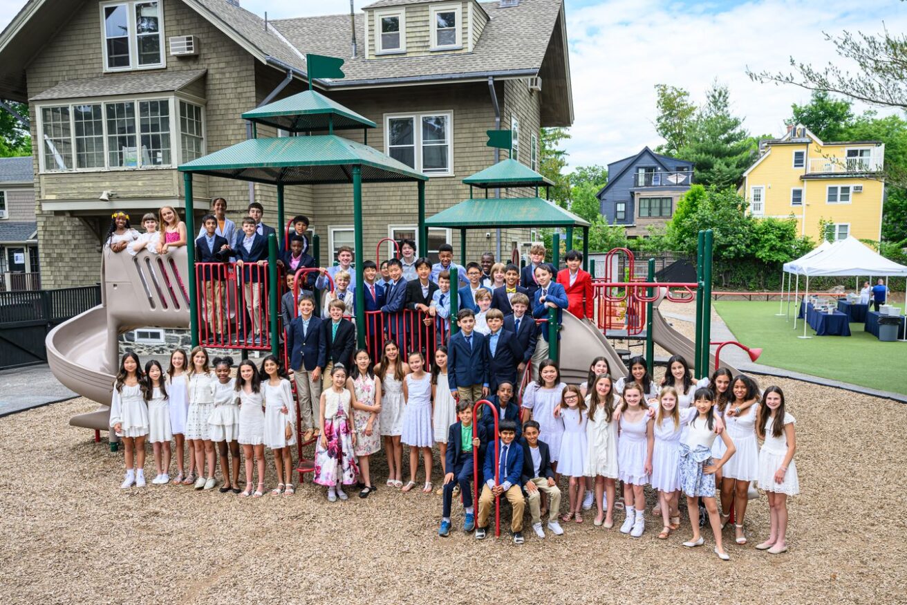 A large group of children, mostly dressed in white, poses on playground equipment in front of a residential-style building.