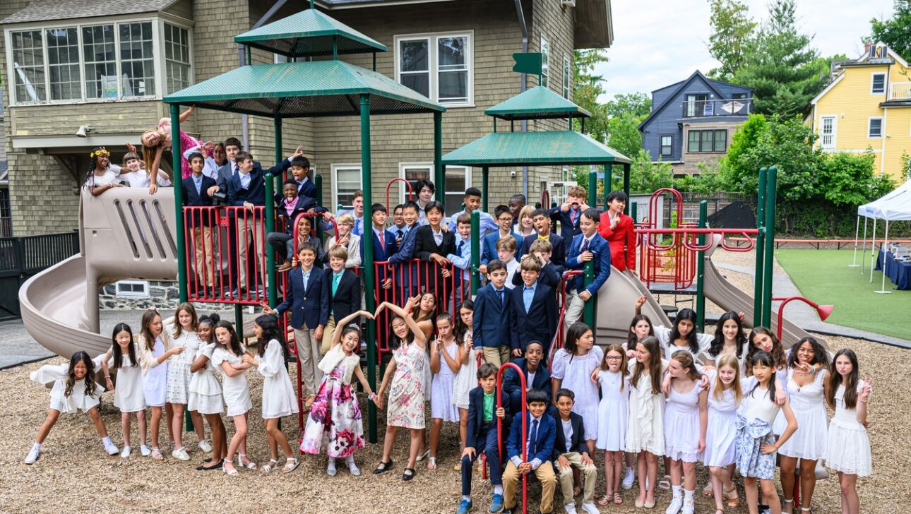 A group photo of approximately 60 children, dressed in formal attire, posing on and around a playground structure in front of a multi-story building.