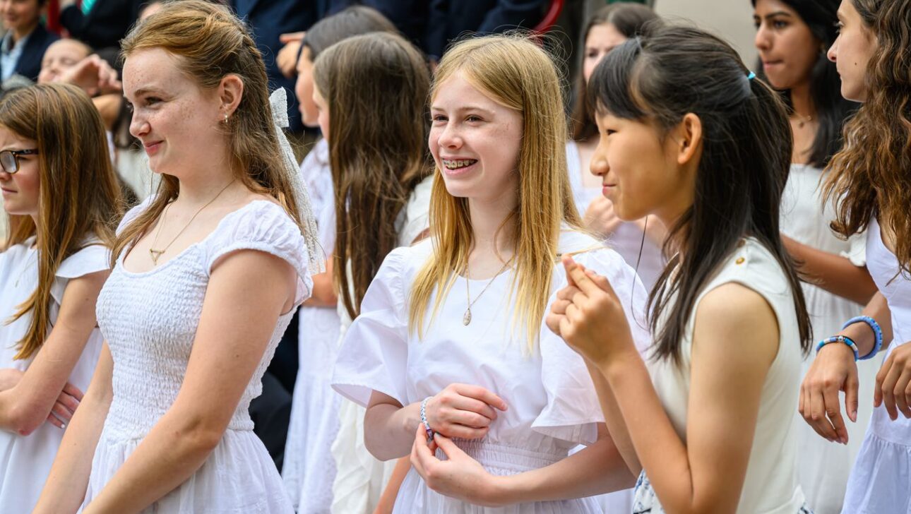 A group of young girls dressed in white dresses are standing together outdoors, smiling and chatting.