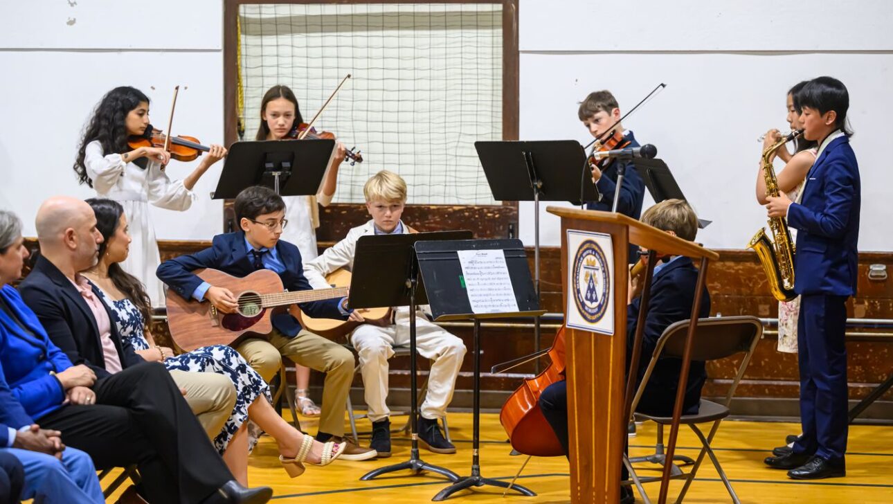 A group of young musicians, including violinists, a guitarist, a cellist, and a saxophonist, performs on a stage at a school event.