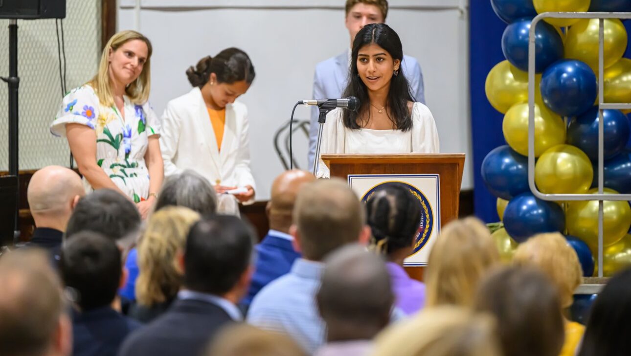 A young woman speaks at a podium in front of a crowd, with three individuals standing behind her and a balloon arrangement on the right.