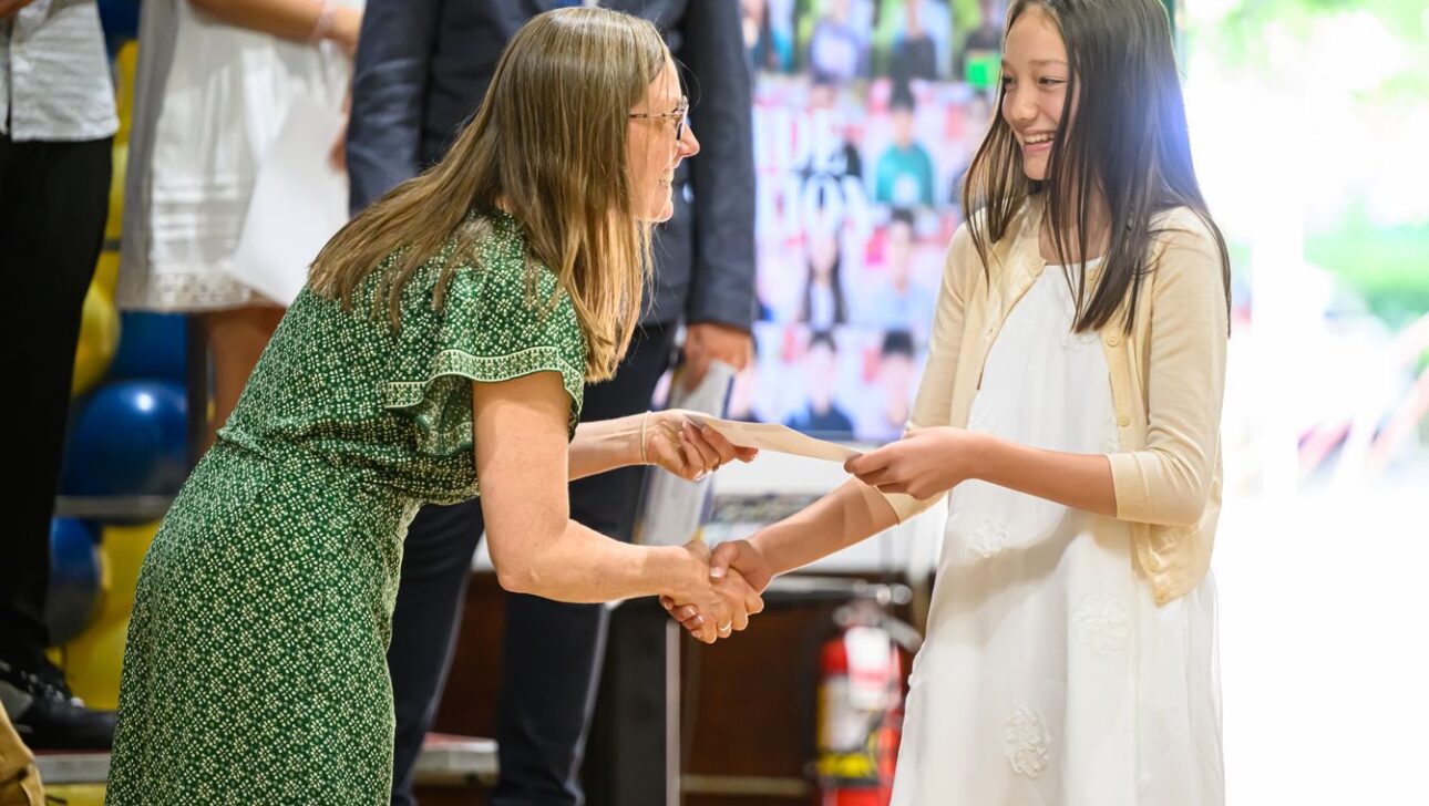 A woman in a green dress shakes hands with a smiling girl in a white dress while handing her a certificate or paper in an indoor setting.