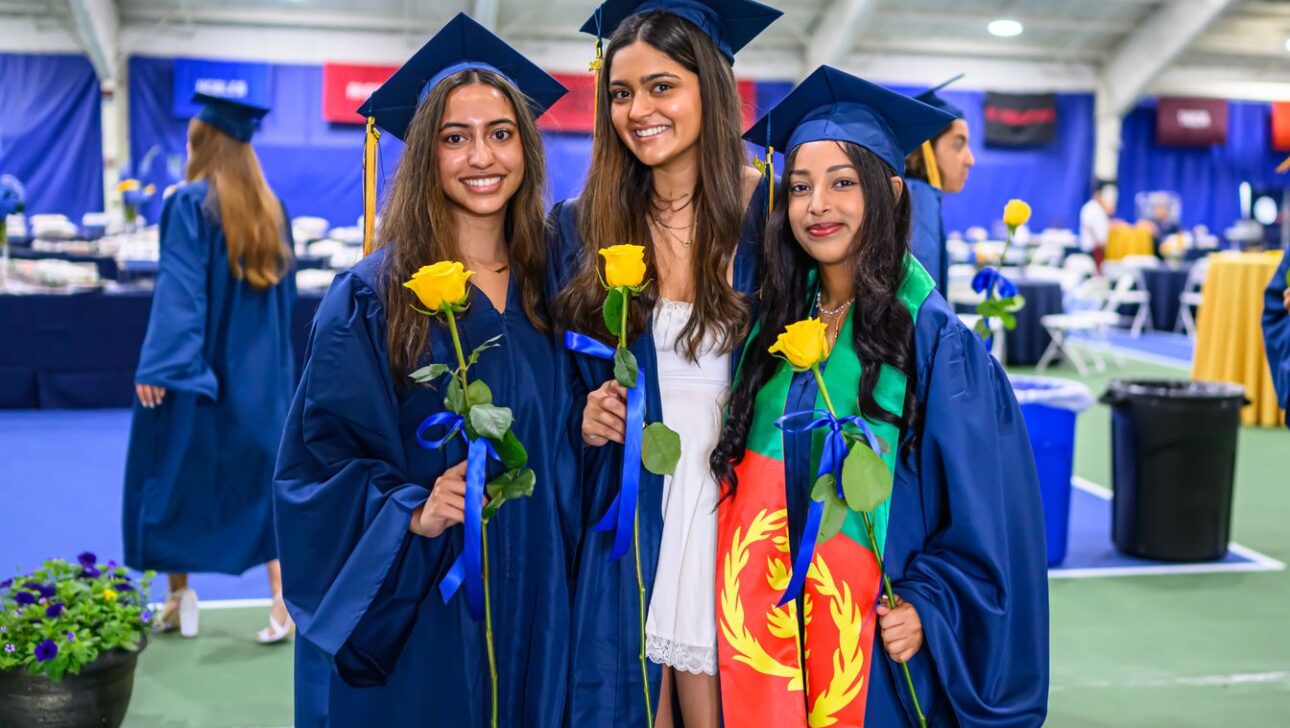 Three students in blue graduation gowns and caps pose for a photo, each holding a yellow rose. One student is wrapped in a flag.