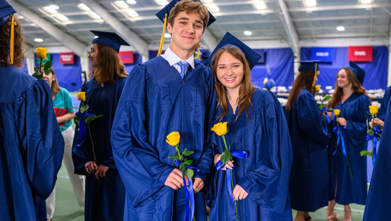 Graduates dressed in blue gowns and caps stand together, holding yellow roses and smiling, at a graduation ceremony indoors.