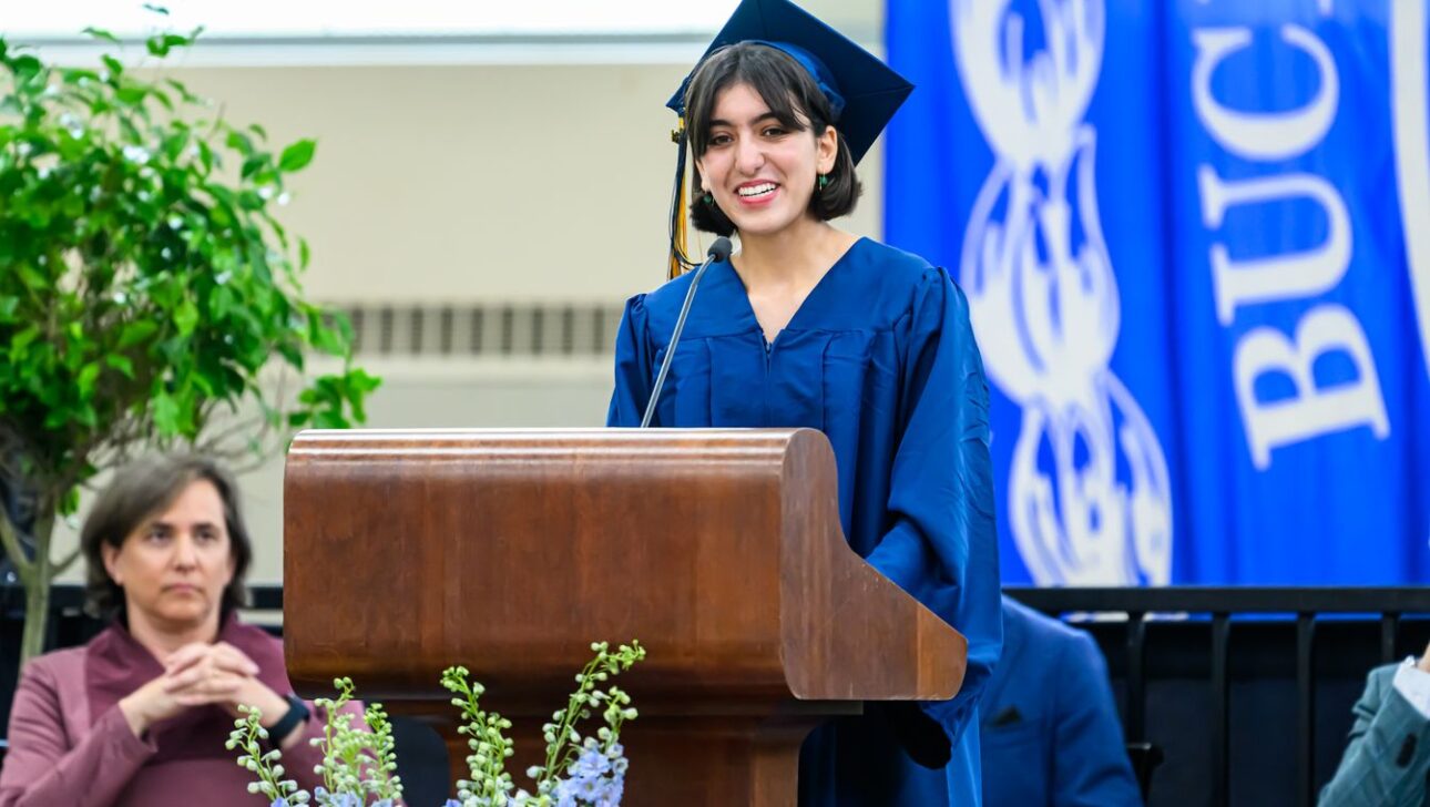 A BB&N graduate in a blue cap and gown speaks at a podium during a ceremony, with an audience member and a banner in the background.