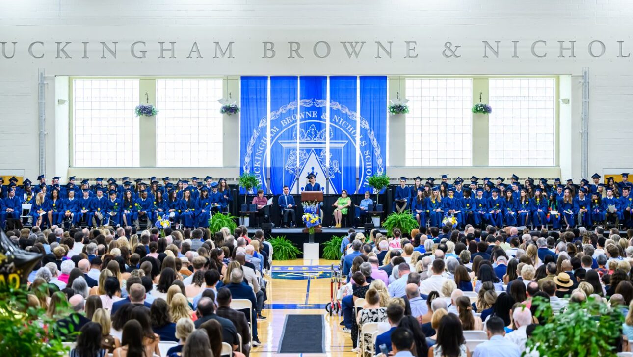 A large group of graduates in blue gowns and caps sit on stage during a commencement ceremony, with an audience seated in front of them.